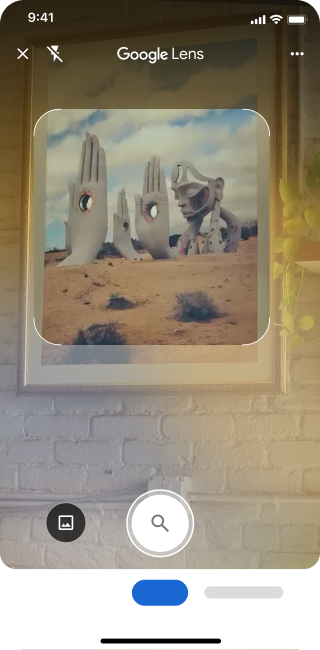 Sur un téléphone, l'application Google Lens est ouverte. Une notification indique à l'utilisateur qu'il peut appuyer sur le bouton de l'obturateur pour rechercher une image de grandes sculptures de mains en pierre dans un désert.